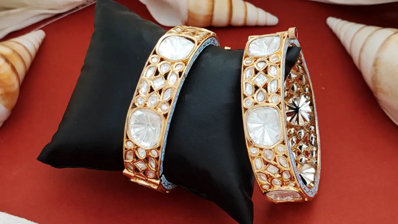 Two gold-tone bangles with clear stones standing on a small black cushion against a red cloth, with shells around them